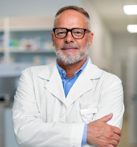 Profissional de saúde grisalho, de barba e óculos, com jaleco branco sobre camisa azul e braços cruzados, em ambiente clínico com prateleiras desfocadas ao fundo, representando a contabilidade para médicos e dentistas