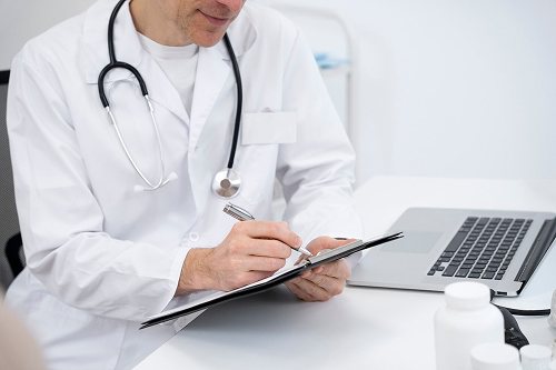 Profissional de jaleco branco com estetoscópio no pescoço escrevendo em uma prancheta, com notebook e frascos sobre a mesa, em uma rotina de contabilidade para médicos e dentistas