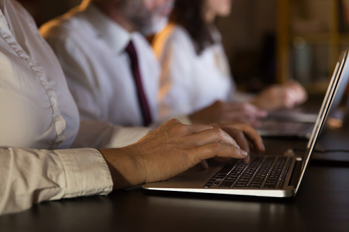 Mãos digitando em um notebook sobre uma mesa escura, com pessoas de camisa social trabalhando em notebooks ao fundo, em uma rotina de contabilidade para startups e infoprodutores